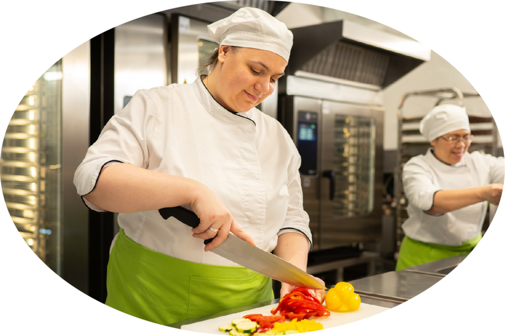 Zwei Köchinnen in weißer Arbeitskleidung und grünen Schürzen in einer Küche.