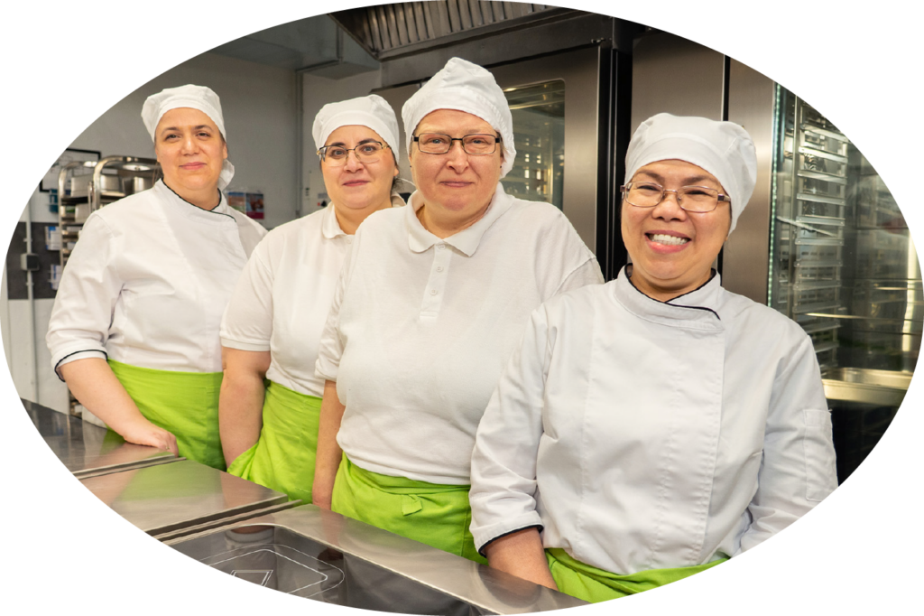 Vier lächelnde Köchinnen in weißer Arbeitskleidung und hellgrünen Schürzen stehen gemeinsam in einer Küche